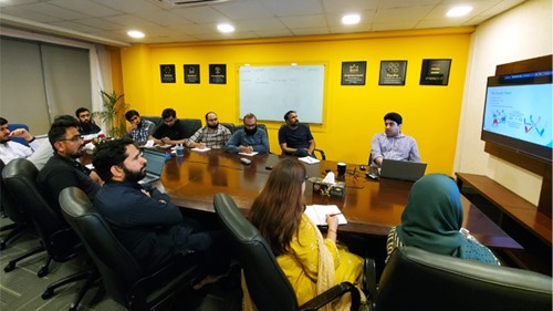 A meeting room is full of people. One person is presenting on a big screen while the rest are listening to that person.