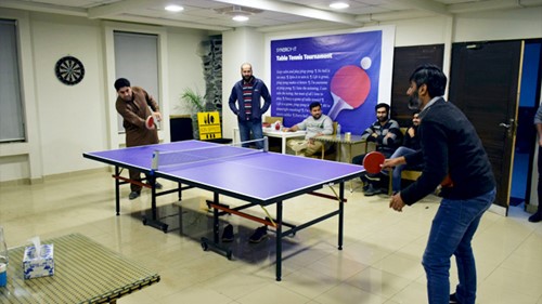 A table tennis tournament is going on. Two people are playing the table tennis game while the rest are looking.
