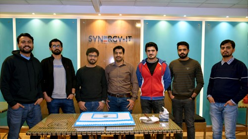 A group of people is standing behind a desk. A cake is placed in the desk.
