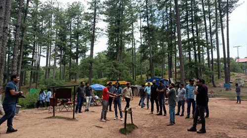 A group of people is standing in a camping site, with tall, green trees in the background.